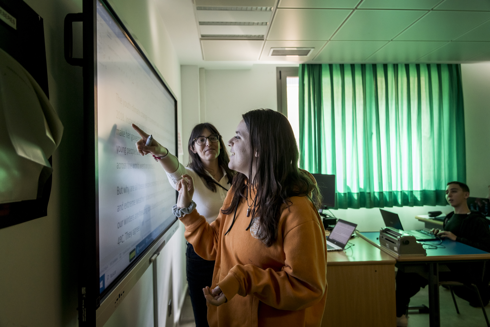 Alumna en un aula señalando un texto en inglés en una pantalla táctil junto a su profesora