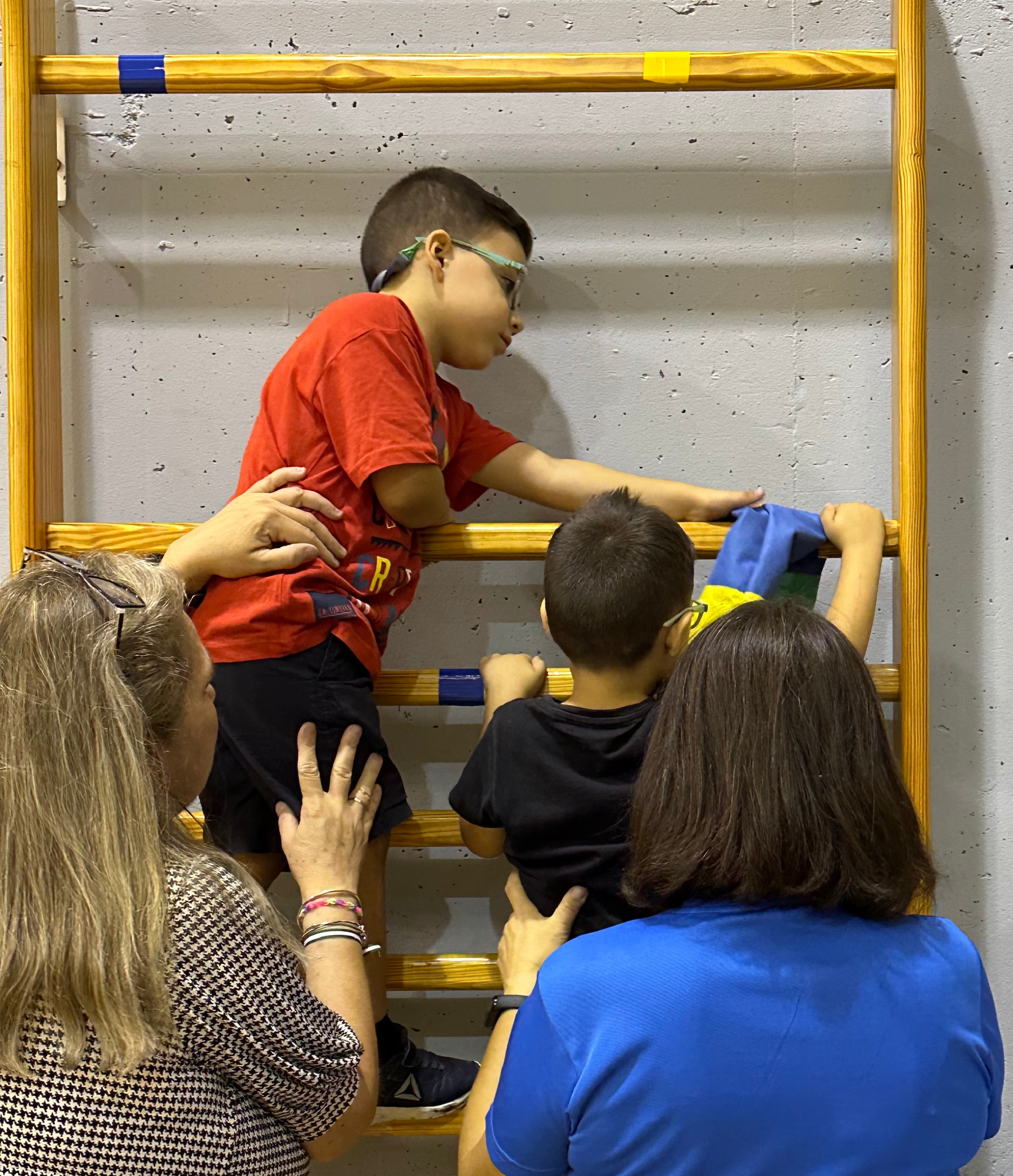 Dos alumnos escalan por unas espaldares bajo el atento cuidado de sus educadoras.