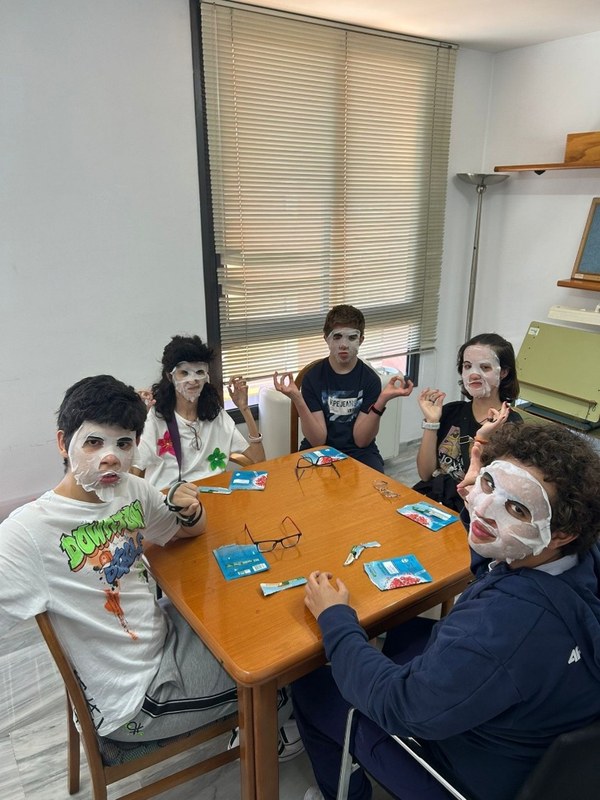 Actividad autocuidado en Murcia Fotografía de un grupo de cinco jóvenes sentados alrededor de una mesa de madera en habitación con ventana y persianas, todos con mascarillas faciales blancas puestas. Sobre mesa, paquetes de mascarillas y gafas, mientras algunos jóvenes hacen gesto de meditación con manos.