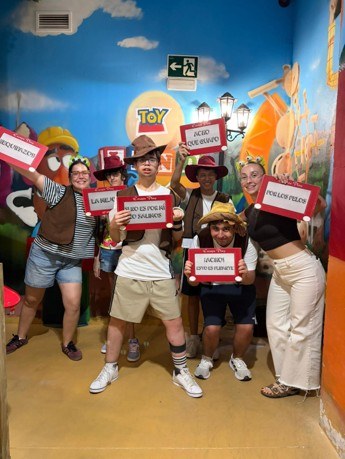 Scape Room en Murcia Fotografía de grupo en sala temática de Toy Story con seis personas posando y sosteniendo carteles con mensajes en español. Fondo colorido con personajes y elementos de la película, todos llevan sombreros de vaquero, transmitiendo ambiente divertido y casual.