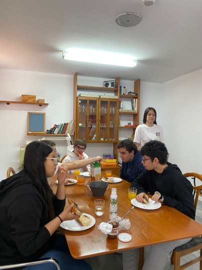 Taller de cocina para adolescentes en Murcia Fotografía de un grupo de cinco adolescentes compartiendo comida y bebida alrededor de una mesa de madera en comedor con estanterías y armario de cristal al fondo. Cuatro personas están sentadas comiendo, una sirve bebida y otra está de pie observando, mostrando ambiente informal y social.