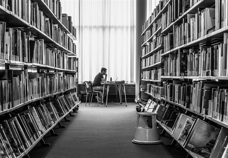 Imagen de una persona estudiando con un ordenador en una biblioteca rodeado de estanterías con libros