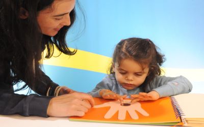 Una niña manipulando la forma de una mano sobre un papel naranja, mientras su madre observa