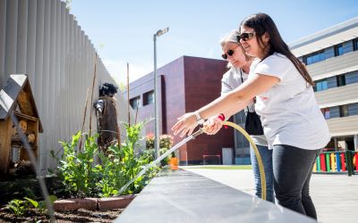 Una alumna regando con una manguera unas plantas en el huerto del centro con ayuda de su maestra
